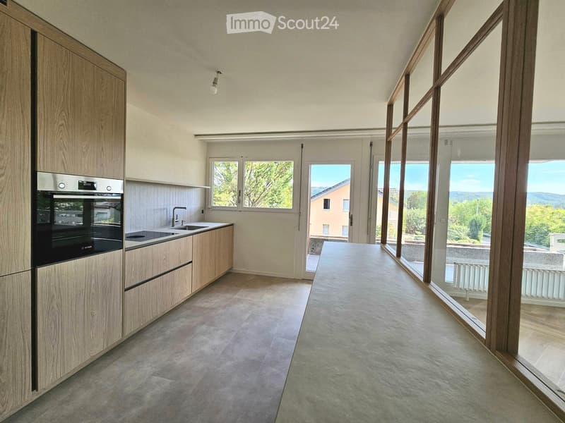 Modern kitchen with light wooden cabinets, stainless steel oven, sink, and large windows offering a scenic view.