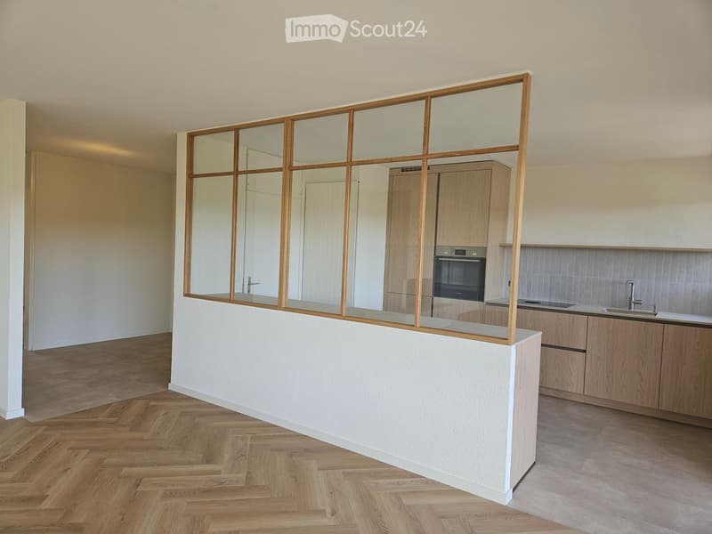 Modern kitchen with light wooden cabinets, a white countertop, and a glass partition with wooden frame separating the kitchen from the living area.