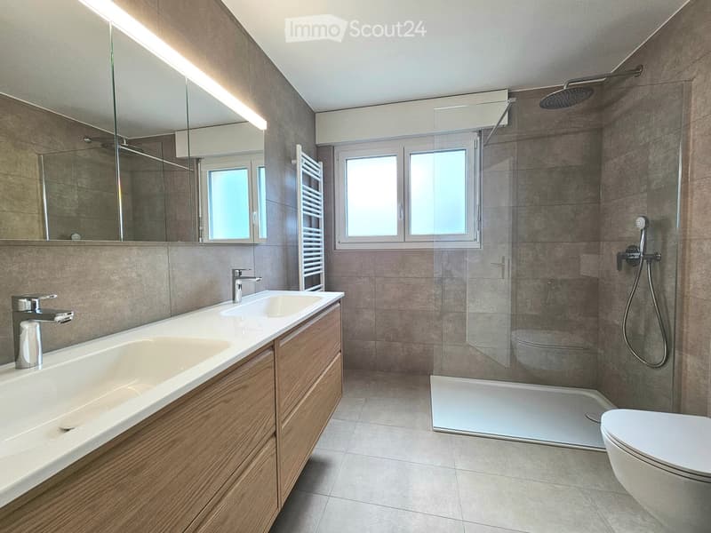 Modern bathroom with a large bathtub, double sink, shower, and toilet. Features wooden vanity, tiled walls, and large windows.