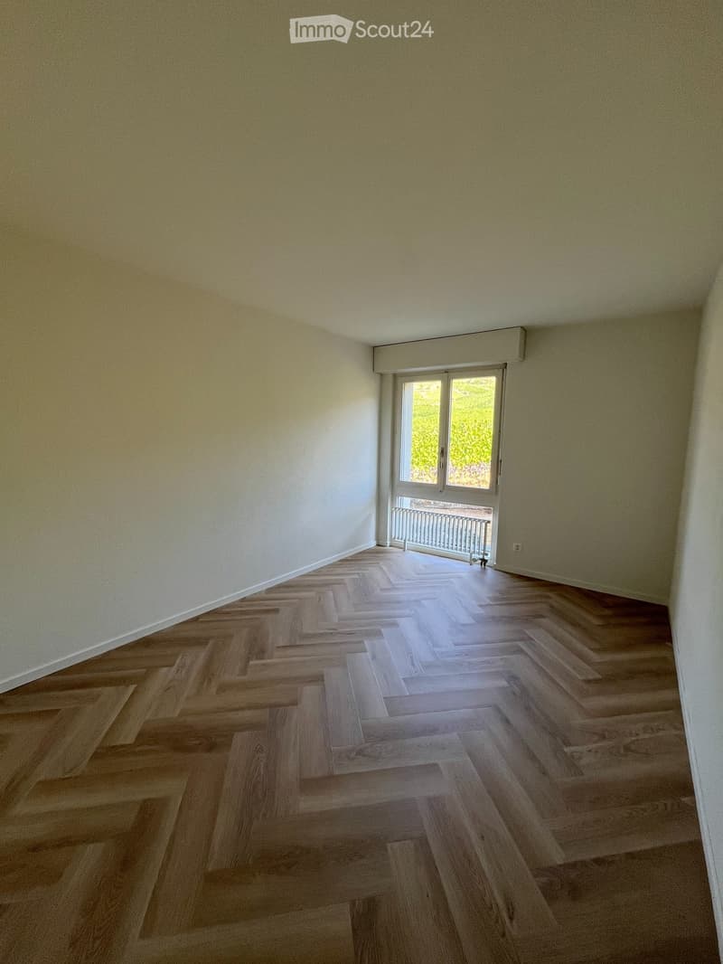 Empty room with light-colored walls and herringbone-patterned wooden flooring, featuring a large window with a view of greenery.