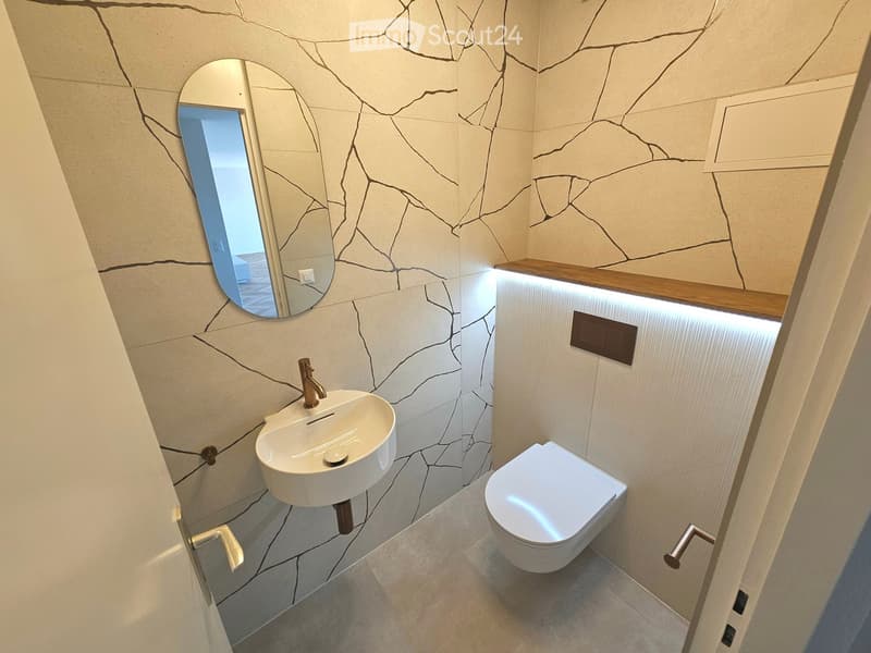 Modern bathroom with a white sink, chrome faucet, toilet, and a round mirror with a wooden shelf above it. The walls have a textured, cracked stone design.