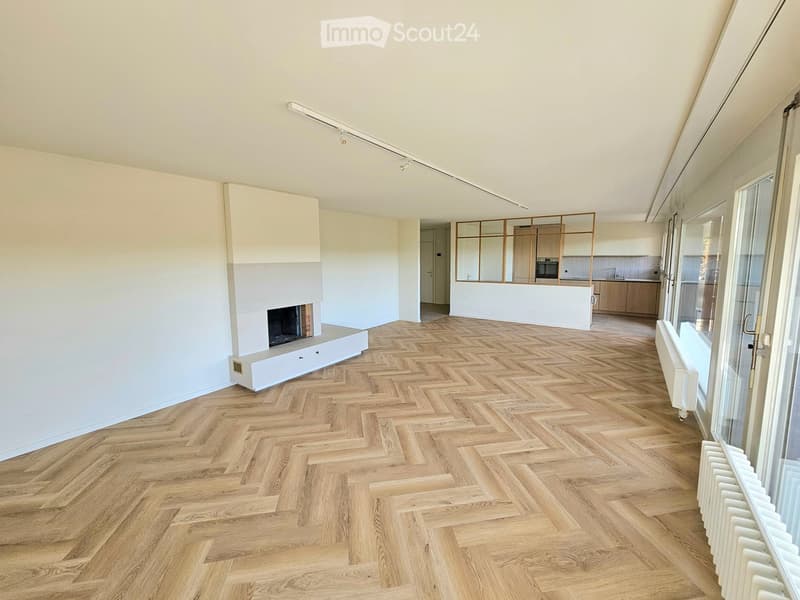 Open-plan living and dining area with herringbone wooden flooring, white fireplace, large windows, and a modern kitchen in the background.