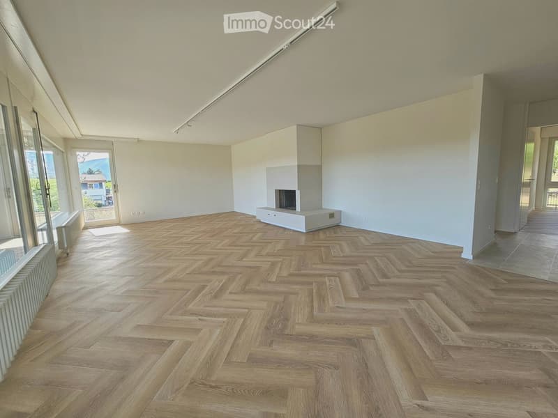 Empty room with herringbone wooden flooring, white walls, large windows, and a built-in fireplace with a white surround.