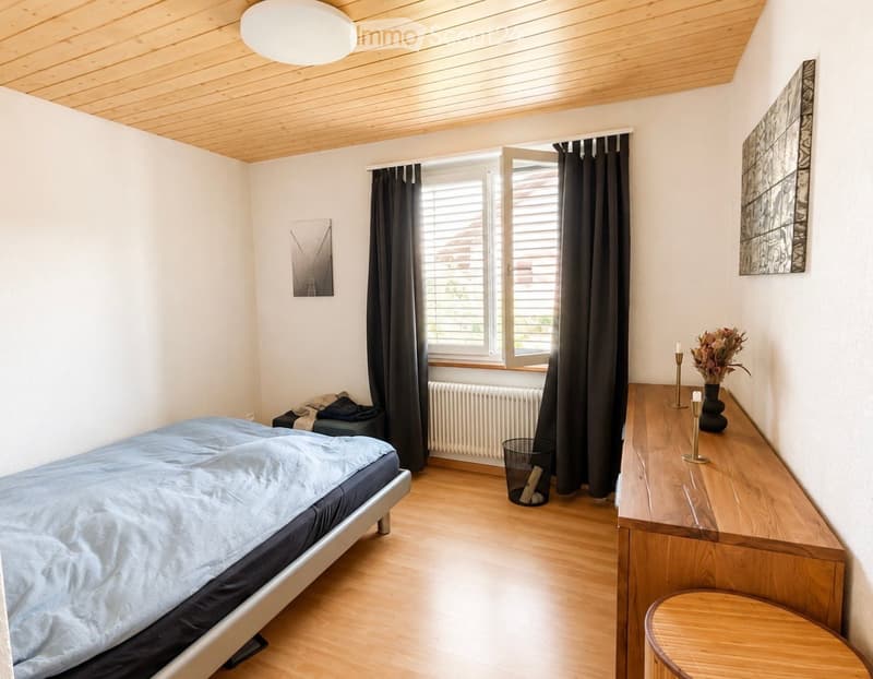 A modern bedroom with a wooden floor, a bed with blue bedding, a nightstand with a vase and candles, a small stool, and artwork on the walls. The room features large windows with dark curtains and a radiator below them.