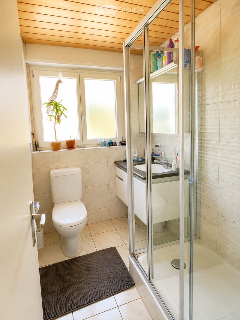 Modern bathroom with a toilet, sink, shower, and a small window with a plant on the windowsill. The space features light-colored tiles and a wooden ceiling.