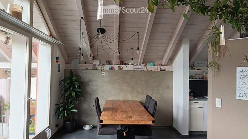 Modern, open-plan room with a wooden dining table, chairs, kitchen area, pendant lights, and plants.