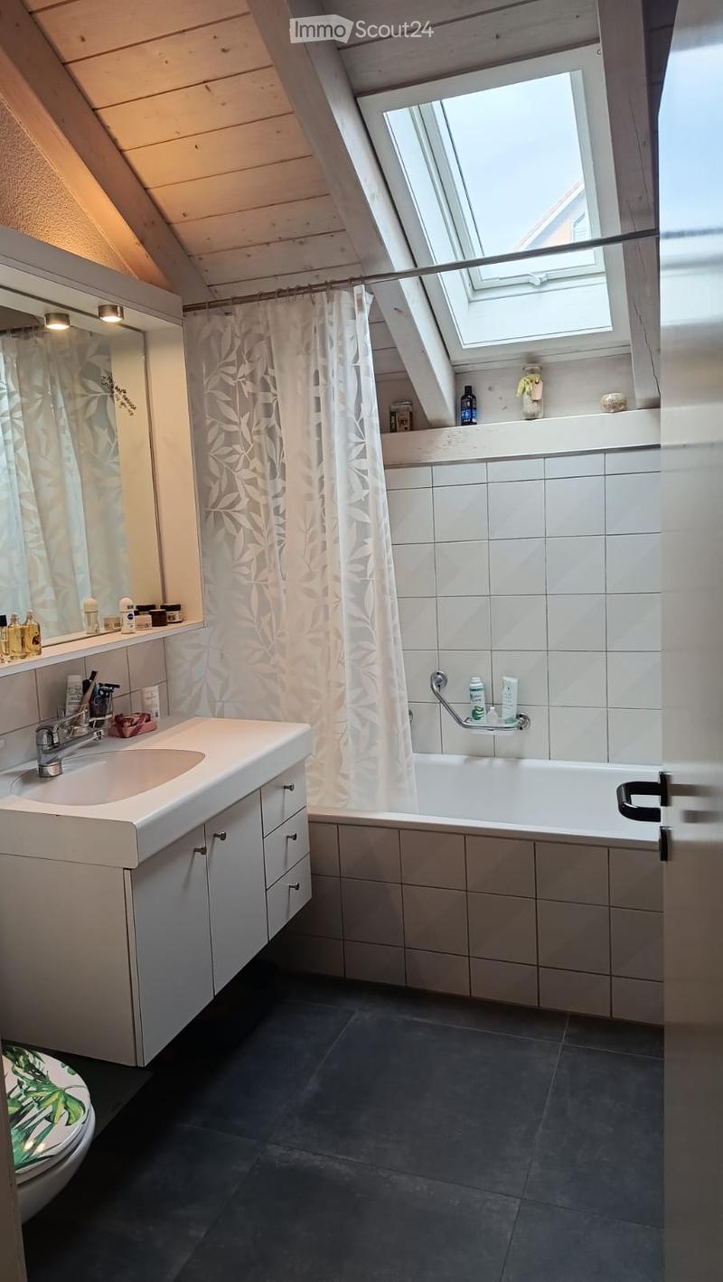 Modern bathroom with a white sink, mirror, bathtub, and toilet. Features a skylight window, dark tiled floor, and various toiletries on the sink and bathtub ledge.
