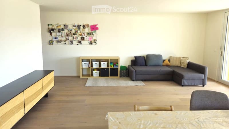 Modern living room with light wooden flooring, grey sectional sofa, coffee table, storage unit, and a dining table with chairs.