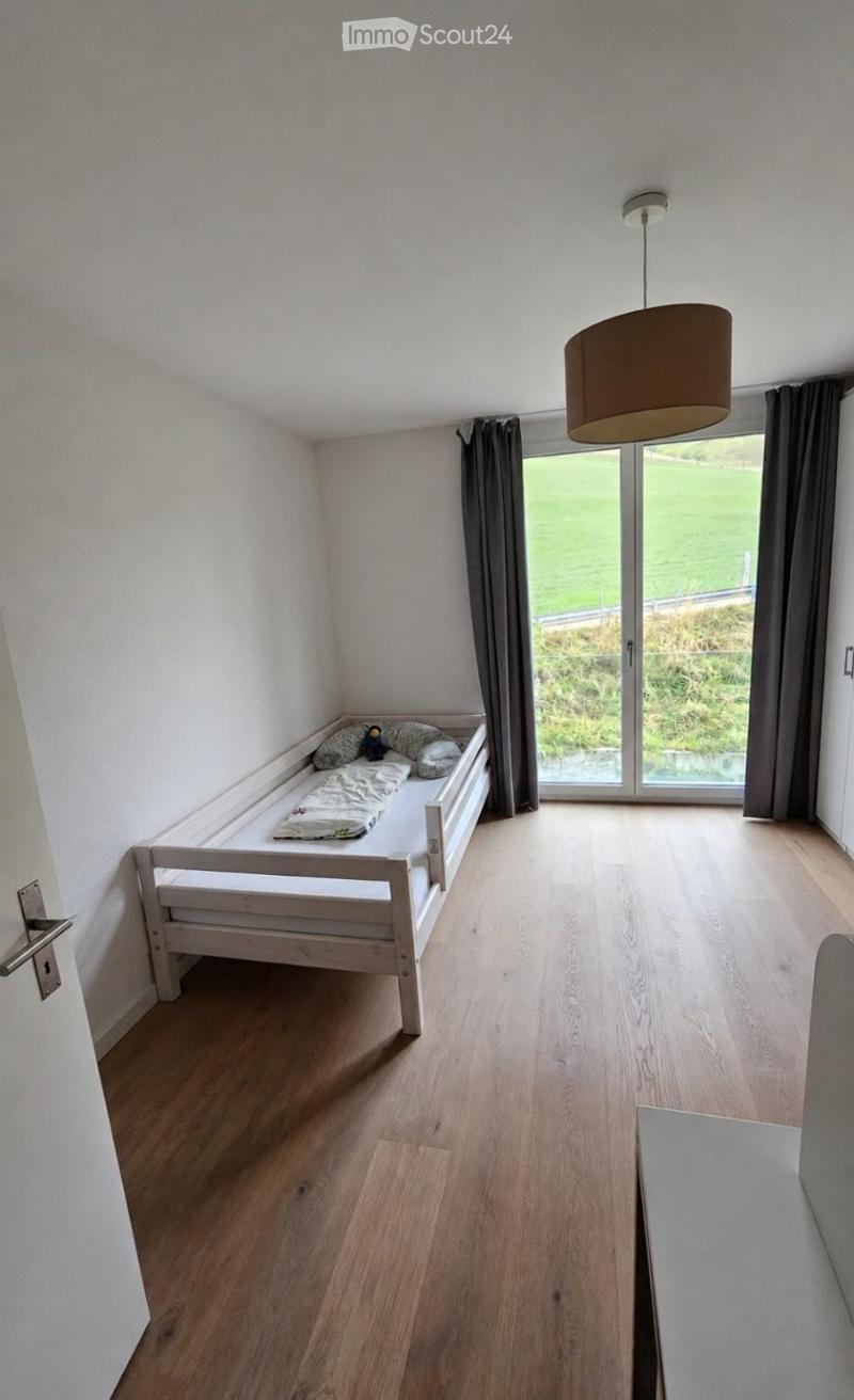 Cozy bedroom with wooden floor, white walls, a single bed with pillows and a stuffed toy, large window with dark curtains, and a hanging lamp.