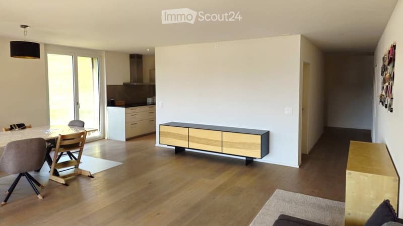 Open-plan living and dining area with light wooden flooring, a modern dining table with chairs, a sleek cabinet, and a contemporary kitchen area with cabinets and a pendant light.