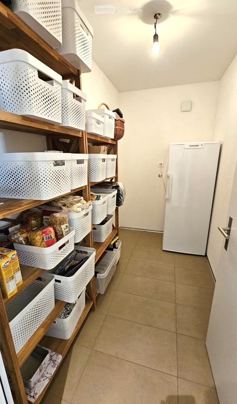A narrow hallway with tiled flooring and white walls. A wooden shelf unit on the left holds multiple white plastic storage bins, some containing food items. A white refrigerator is positioned against the right wall.
