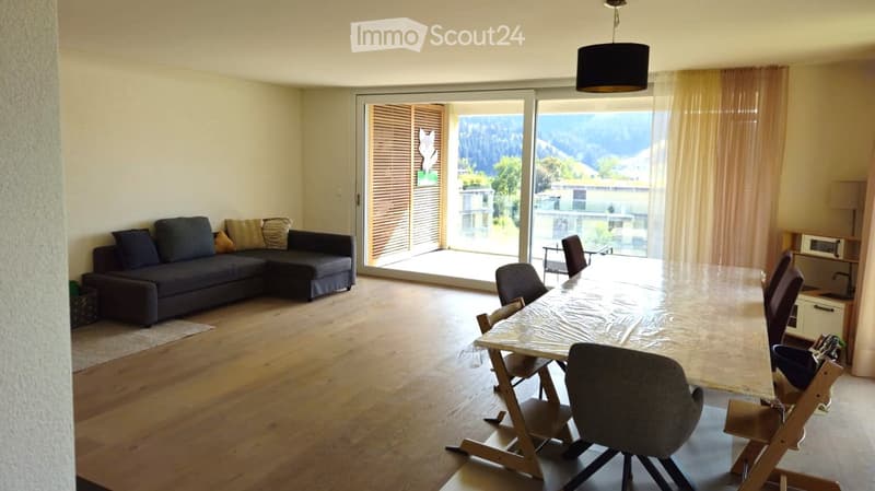 Bright living and dining area with wooden flooring, large windows offering a scenic mountain view, a dark-colored sectional sofa, a long dining table with chairs, and a hanging pendant light.