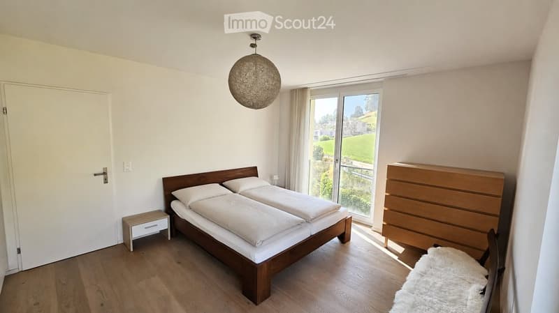 Bright bedroom with wooden flooring, a double bed with white bedding, a nightstand, a wooden dresser, and a chair with a white rug.