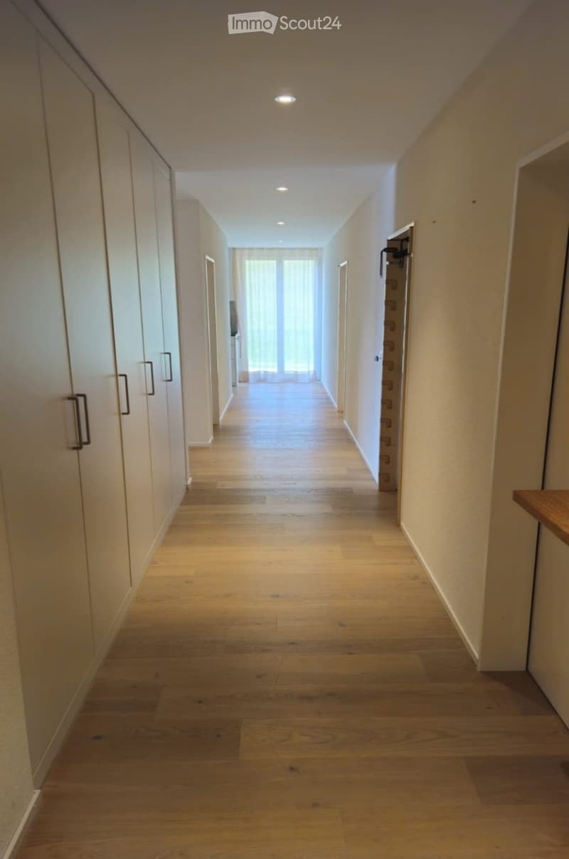 A long, well-lit hallway with wooden flooring, white walls, and built-in wardrobes on one side. Natural light streams in from a window at the end of the corridor.