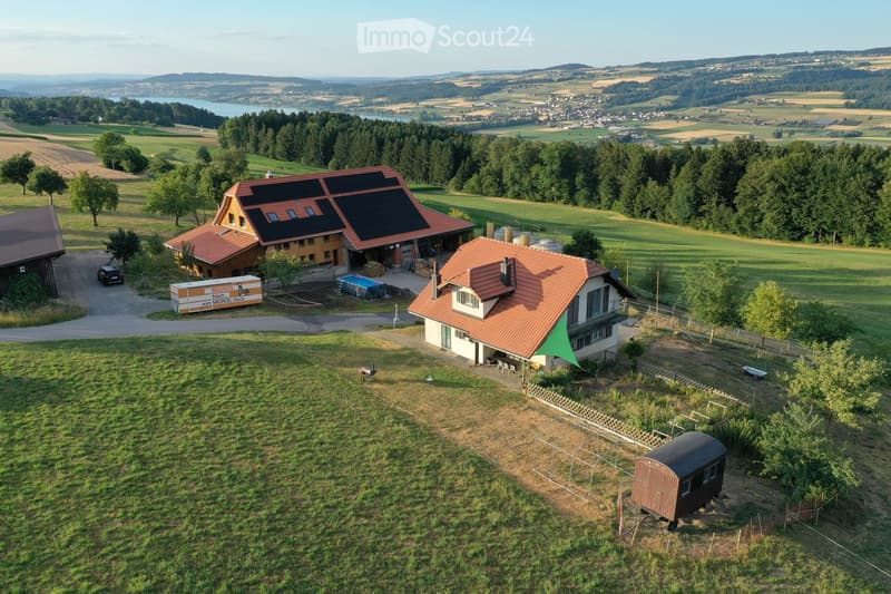 Aerial view of a rural property with multiple buildings, including a main house with a red-tiled roof, a smaller house, a garage, and a small shed. The property is surrounded by green fields, trees, and a scenic landscape with hills and a lake in the back