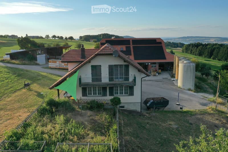 A two-story house with a red-tiled roof, surrounded by a well-maintained garden and open fields. The house features large windows, a balcony, and solar panels on the roof. There is a barbecue, a green umbrella, and a fenced garden area visible.