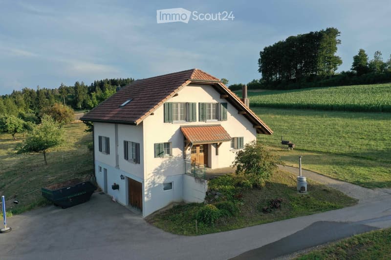 Single-story house with a red-tiled roof, white exterior, multiple windows with shutters, and a small porch with a wooden door. The house is surrounded by a grassy field with trees and a cornfield in the background.