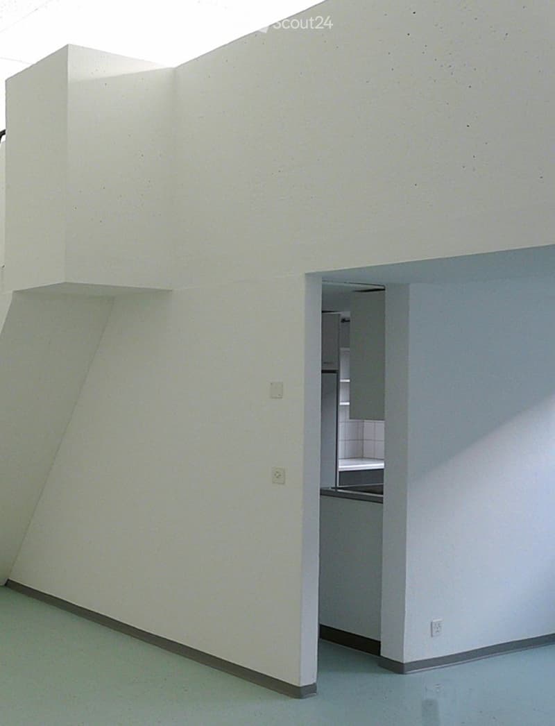 Empty room with white walls and light blue flooring, featuring an open doorway leading to a small kitchen area with white cabinets and a sink.