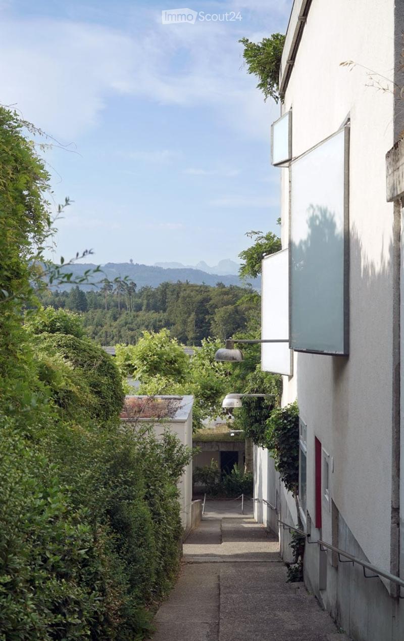 A narrow, tree-lined pathway between two modern buildings, leading towards a distant view of mountains and greenery.
