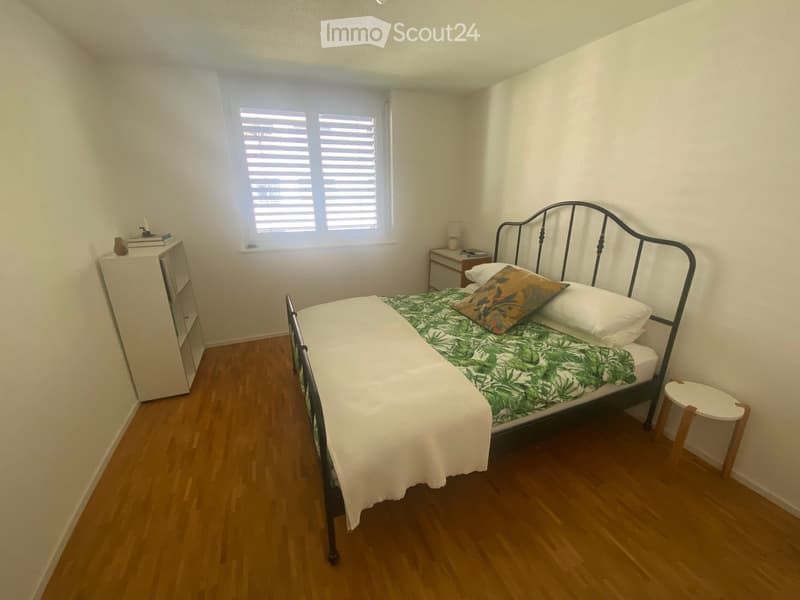Cozy bedroom with wooden flooring, a single bed with a green and white patterned bedspread, a nightstand with a lamp, a small stool, and a white wardrobe.