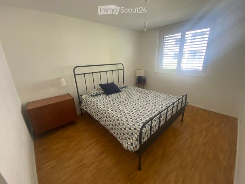 A well-lit bedroom with wooden flooring, a metal-framed bed with a patterned duvet, two nightstands with lamps, and a small wooden drawer unit.