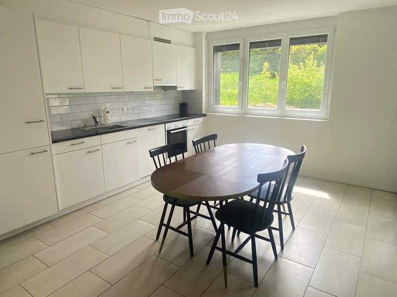 Modern kitchen with white cabinets, a round wooden dining table, and four chairs. The kitchen features a black countertop, a sink, and an oven, with large windows offering a view of greenery.