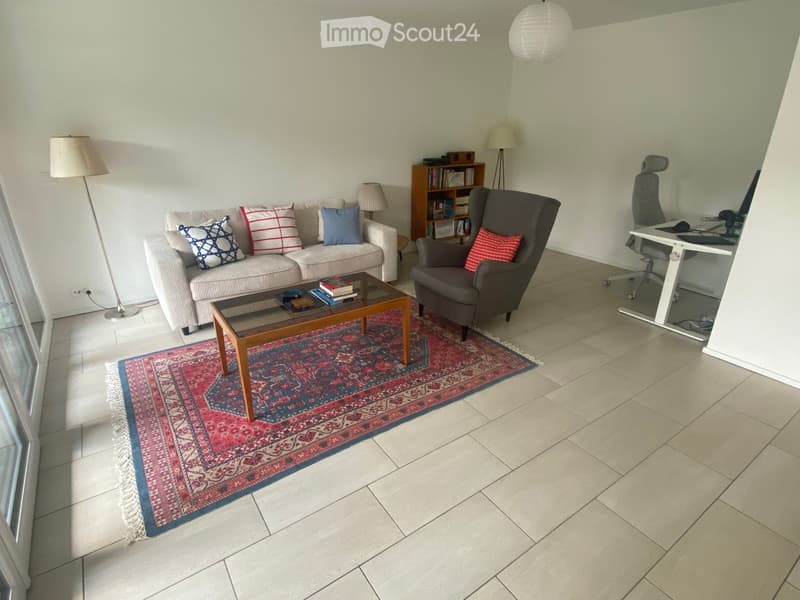 A modern living room with a sofa, coffee table, armchair, desk, and bookshelf. The room features a large area rug and several lamps.