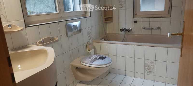 Bathroom with a toilet, bathtub, sink, and tiled walls. Features a handrail for accessibility and a small window above the bathtub.