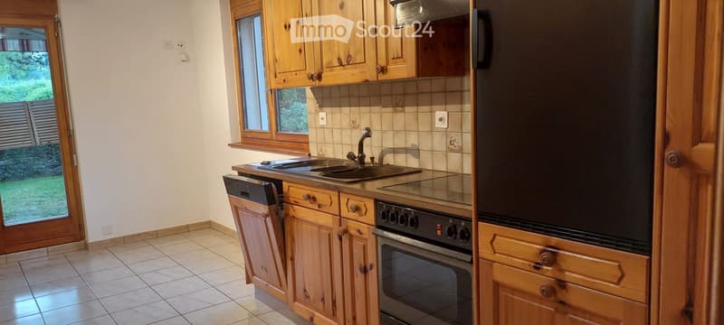 A well-lit kitchen with wooden cabinets, a sink, oven, refrigerator, and dishwasher. The space features tiled flooring and a window with a view of the garden.