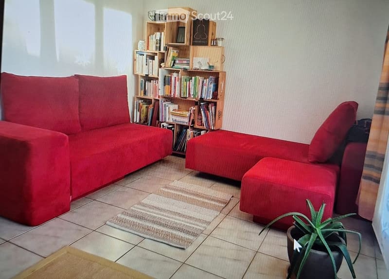 A cozy living room with a red sectional sofa, a matching red ottoman, a wooden bookshelf filled with books, a small potted plant, and a patterned rug on the tiled floor.