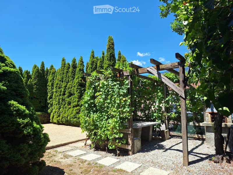 Well-maintained garden with neatly trimmed coniferous trees, a wooden pergola, a stone table, and a gravel pathway.