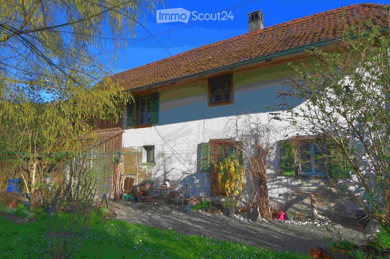 Traditional house with red roof, white walls, green shutters, garden with plants and trees, outdoor seating area with table and chairs