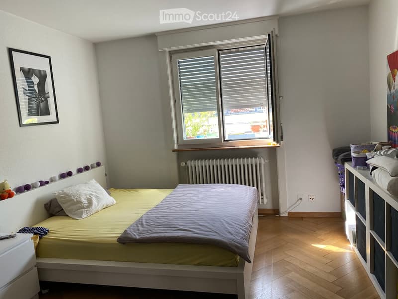 Cozy bedroom with a single bed, nightstand, radiator, and a window with blinds. The room features wooden flooring and a picture frame on the wall.