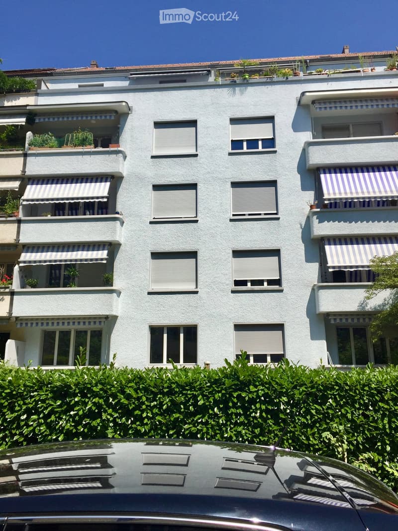 Four-story residential building with white facade, multiple windows, balconies with striped awnings, and rooftop greenery.