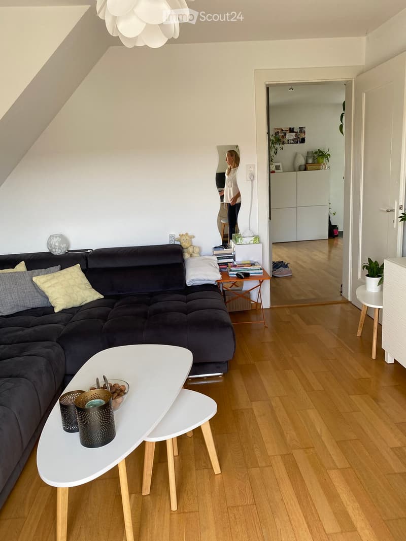 Modern living room with wooden flooring, a dark grey sectional sofa, a white coffee table, side table, and a potted plant. The room features a slanted ceiling and a mirror reflecting part of the space.