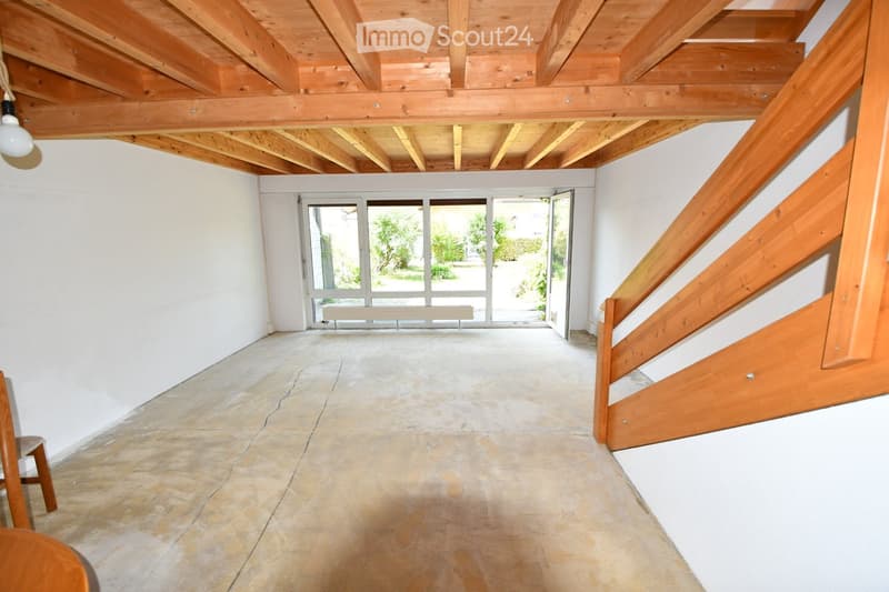 Empty room with exposed wooden beams on the ceiling, a staircase with wooden railings, a large window with a bench, and a small wooden chair near the wall.