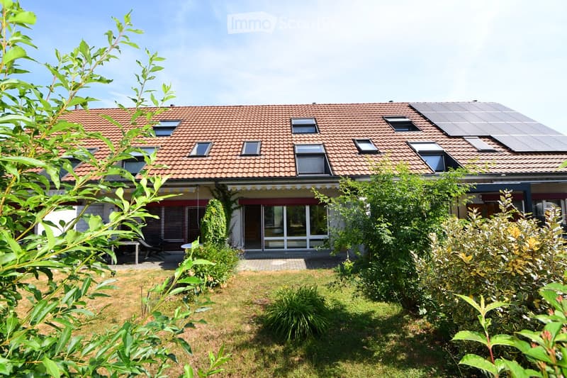 Two-story house with red-tiled roof, multiple windows, solar panels on the roof, and a well-maintained garden with greenery and a patio area.