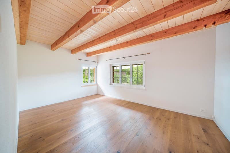 Empty room with wooden flooring, white walls, exposed wooden beams on the ceiling, and two windows with white frames.