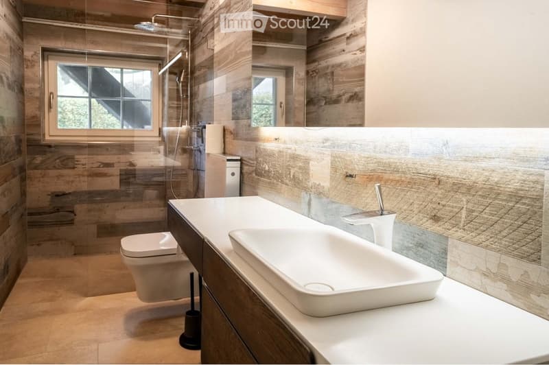Modern bathroom with wooden accents, a large sink with a sleek faucet, a toilet, and a glass-enclosed shower area with a modern showerhead.