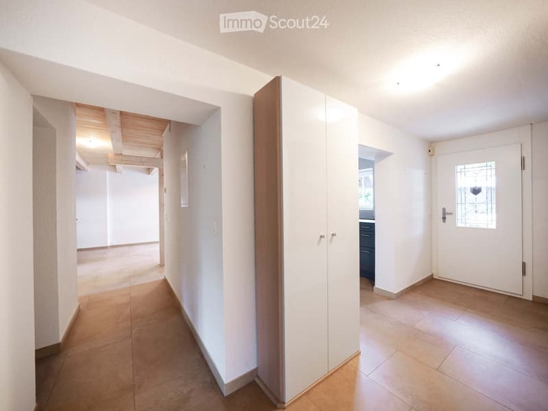 Bright hallway with tiled flooring, white walls, and built-in white closet with wooden trim. Features a wooden ceiling in the background and a door leading to another room.