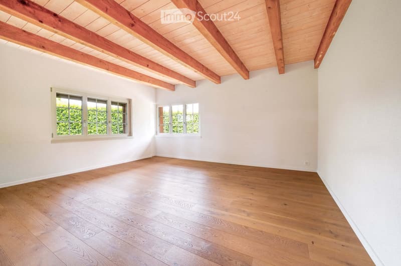 Empty room with wooden flooring, white walls, and exposed wooden beams on the ceiling. Two windows with white frames are present, allowing natural light to enter.