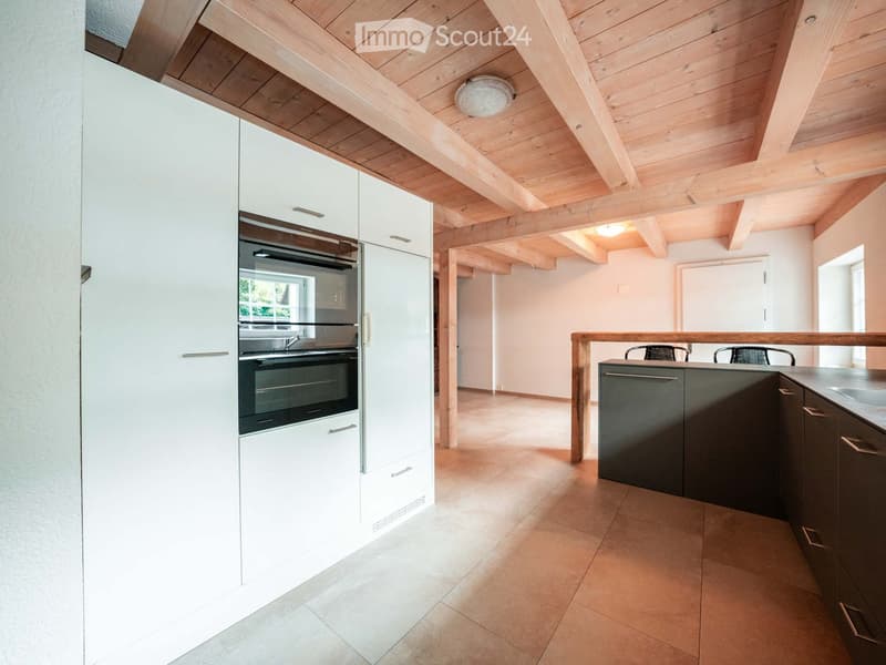 Modern kitchen with white and dark grey cabinets, an integrated oven, and a wooden ceiling with exposed beams. The space features a tiled floor and a small seating area with chairs.