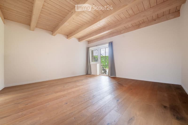 Empty room with wooden flooring, white walls, and a wooden ceiling. Features a large window with grey curtains and a sliding glass door leading outside.