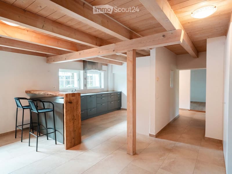Modern kitchen with exposed wooden beams, bar counter, stools, and grey cabinets.