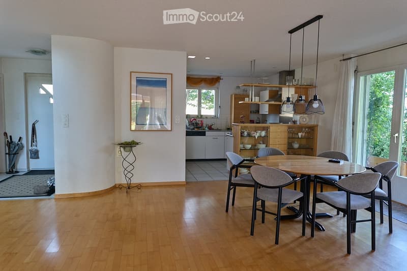 The image shows a modern, well-lit living and dining area with light wooden flooring. A round wooden dining table with six matching chairs is positioned in the center. The kitchen area features white cabinets, various appliances, and a countertop with ite