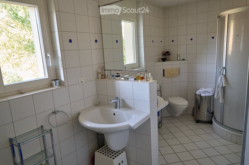 Bright bathroom with white tiled walls and floor, featuring a sink with a modern faucet, a large mirror, a toilet, a shower, and a glass shelf holding various toiletries.