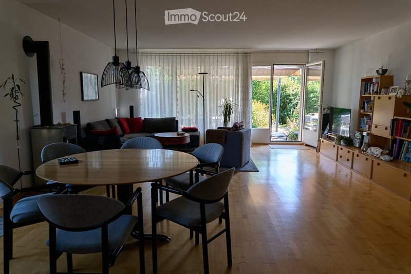 Bright living room with wooden flooring, large windows, and a balcony. Features a sofa, coffee table, dining table with chairs, bookshelf, and television.