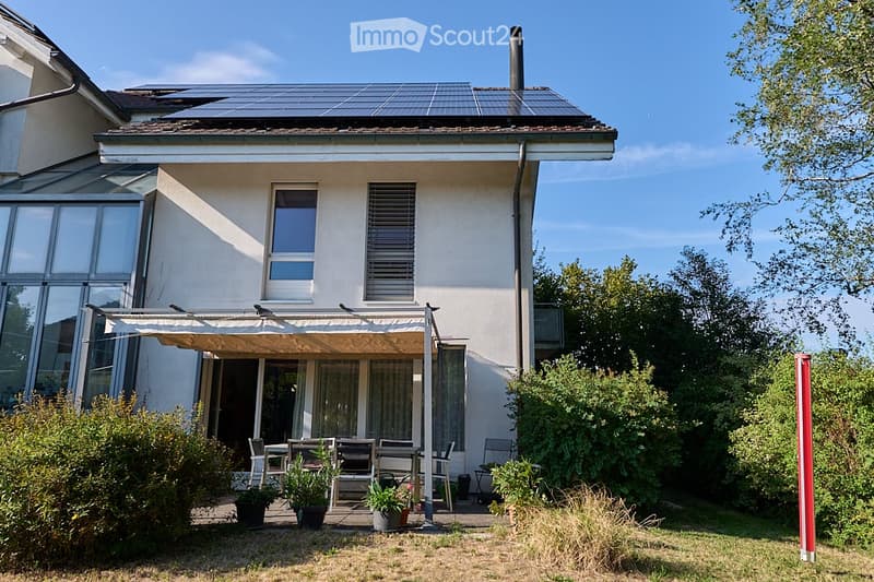 Two-story house with solar panels on the roof, large windows, and a covered patio area with outdoor furniture.