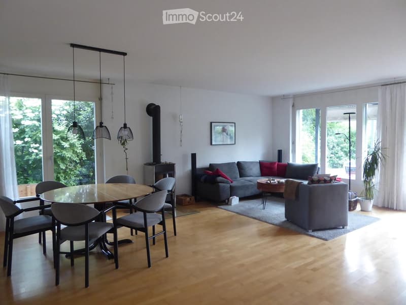 Bright living room with wooden flooring, large windows, grey sofa, coffee table, dining table with chairs, and a fireplace.