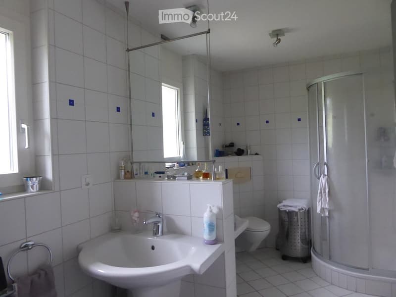 Modern bathroom with white tiles, a sink, a toilet, a shower, and a mirror. The sink has a faucet and a bottle on the counter. A towel hangs on the side of the shower, and a trash can is placed near the toilet.